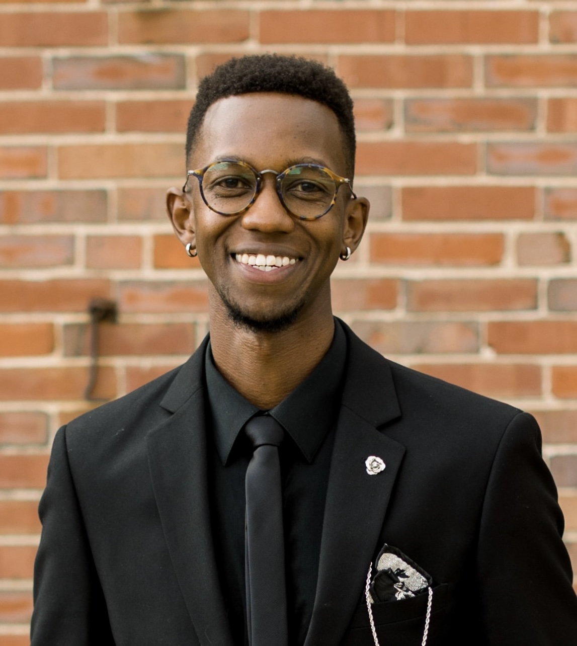 Kip Kiplagat smiling in a dark suit and glasses in front of a brick wall